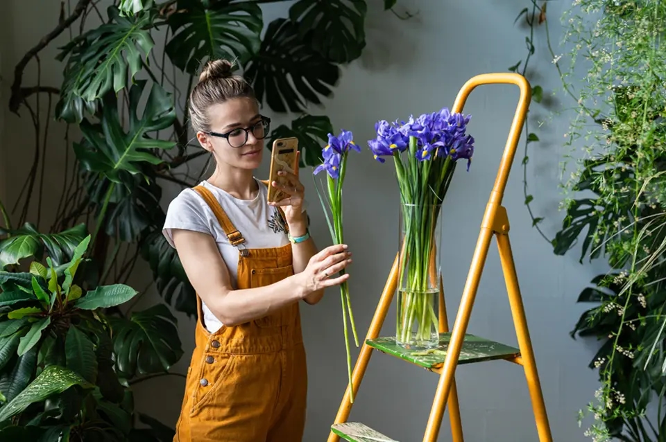Kobieta fotografuje bukiet fioletowych irysów w wazonie, otoczony roślinami doniczkowymi.